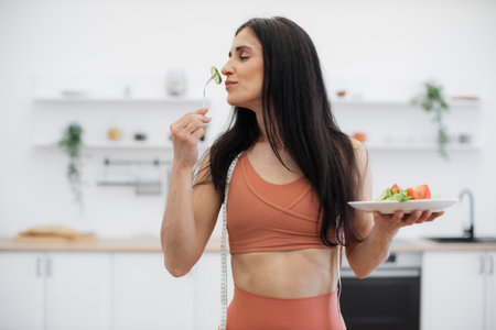 Energetic fitness woman enjoying smell of fresh cucumber while eating low-calorie food after efficient training at home. Active lady taking care of health by consuming organic meal and working out.の写真素材