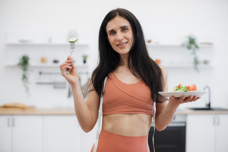 Energetic fitness woman enjoying smell of fresh cucumber while eating low-calorie food after efficient training at home. Active lady taking care of health by consuming organic meal and working out.の写真素材