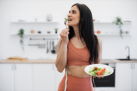 Long-haired brunette looking aside while trying freshly prepared vegan salad in spacious white kitchen on blurred background. Gorgeous woman promoting healthy eating for excellent well-being.の写真素材