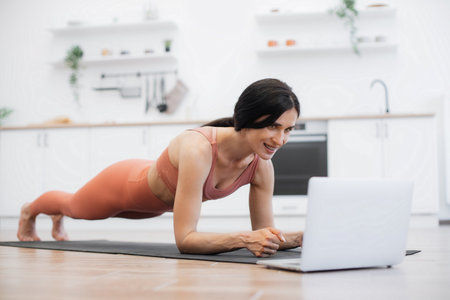 Side view of caucasian fitness woman dressed in activewear doing plank exercise on yoga rug while staying at home. Fit lady listening motivational tracks on remote laptop. Concept of active lifestyle.の写真素材