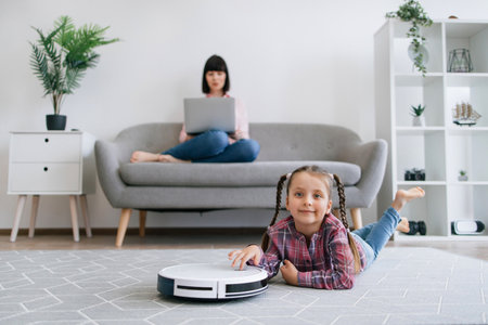 Cute tween girl in casual wear lying on floor near robot vacuum cleaner while busy lady typing on laptop on sofa. Carefree daughter having fun with wireless device during moms working day at home.の写真素材