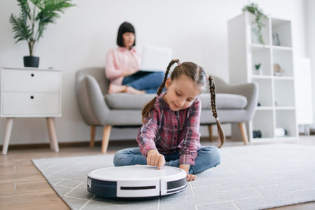 Close up view of little girl pressing button on robotic vacuum while attractive female doing full-time job indoors. Smart electronic equipment helping parents turning home care into joy for kids.の写真素材
