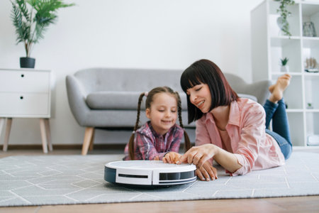 Close up view of joyful woman and tween girl activating robotic cleaner while resting on carpet in apartment. Delighted parent and child following rules of using smart electrical appliance at home.の写真素材