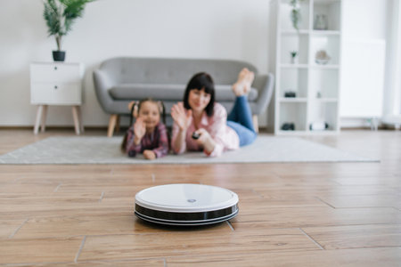 Focus on robot vacuum carrying out scheduled cleaning task while female owners waving goodbye from behind. Smart device saving energy from tedious home cleaning work by using advanced technologies.の写真素材