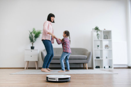 Modern robot vacuum performing automatic cleaning while barefoot woman and girl dancing in background. Happy caucasian family moving to beat of relaxing songs without worries about household chores.の写真素材
