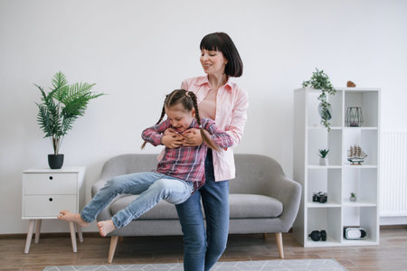 Strong sporty mom twirling preteen child around with hands under armpits while staying at home on sunny day. Emotional caucasian family woman motivating playful daughter to leading active lifestyle.の写真素材