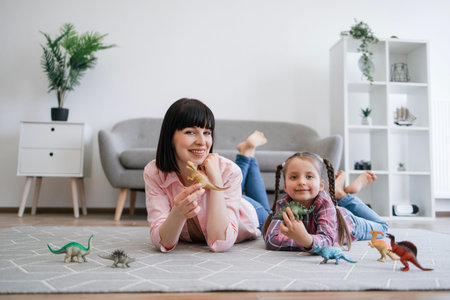 Portrait of mother-daughter duo posing with amazing toys of extinct reptiles in hands while lying comfortably on floor. Smiling caucasian ladies gaining knowledge about different animals and habitats.の写真素材