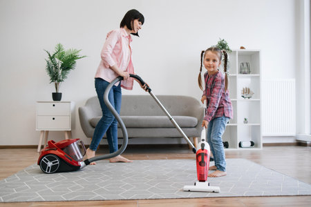 Full length view of preteen girl holding upright vacuum cleaner while female grown-up using canister model alongside. Prudent ladies of house collecting dust on various surfaces at sunny weekend.の写真素材