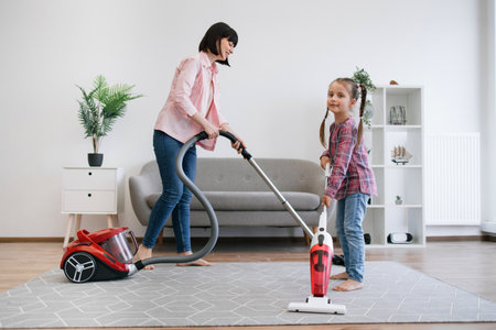 Full length view of preteen girl holding upright vacuum cleaner while female grown-up using canister model alongside. Prudent ladies of house collecting dust on various surfaces at sunny weekend.の写真素材