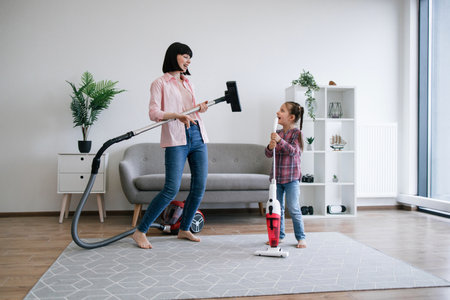Excited mother using vacuum cleaner as imaginary guitar while daughter pretending rock singer with cordless device. Overjoyed ladies of house having fun while sharing home care duties at weekend.の写真素材