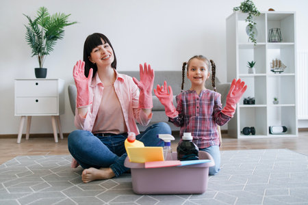 Portrait of smiling mother and daughter showing hands in rubber gloves while sitting near plastic tub with cleaning supplies. Cheerful homemakers getting ready for polishing and dusting off room.の写真素材