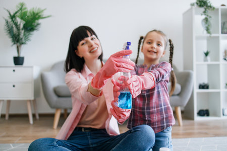 Focus on half-full spray cleaner held by positive caucasian woman and little girl in denim outfits sitting in living room. Multipurpose detergent helping homemakers to fight dirt on various surfaces.の写真素材