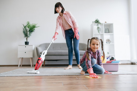 Preteen kid in latex gloves cleaning floor from dirt while mother removing dust from carpet with canister vacuum. Tireless ladies of house reducing risk of allergies by taking care of home chores.の写真素材