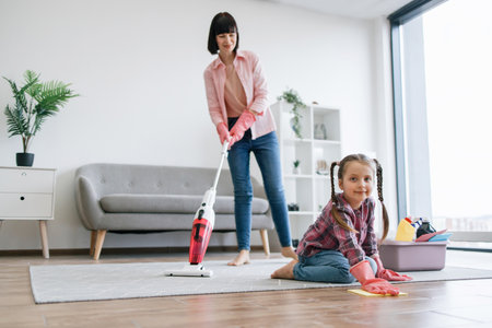 Preteen kid in latex gloves cleaning floor from dirt while mother removing dust from carpet with canister vacuum. Tireless ladies of house reducing risk of allergies by taking care of home chores.の写真素材