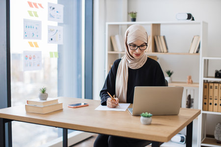Woman in hijab making notes from laptop screen in officeの写真素材