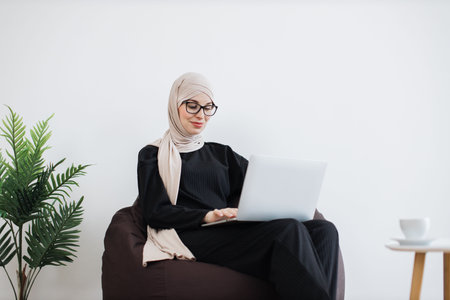 Arabian woman with computer resting in beanbag in workspaceの写真素材