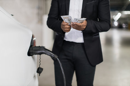 Adult man holding dollar bills near e-car fill-up in parkingの写真素材