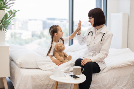 Pediatrician and girl giving high five to each other on bedの写真素材