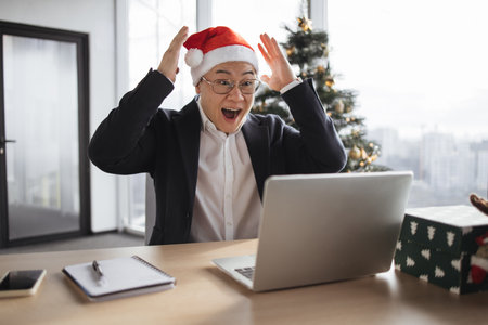 Emotional adult in Santa hat lifting hands while looking at computer screenの写真素材