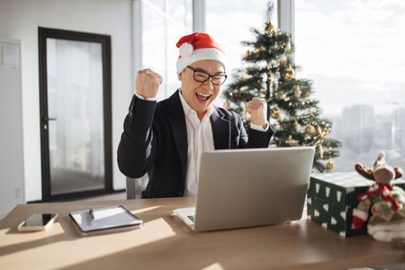Emotional adult in Santa hat lifting hands while looking at computer screenの写真素材