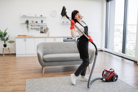 Beautiful woman in black apron vacuuming carpet in bright living room kitchen.の写真素材