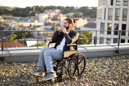 Thoughtful man in wheelchair on rooftop overlooking cityscapeの写真素材