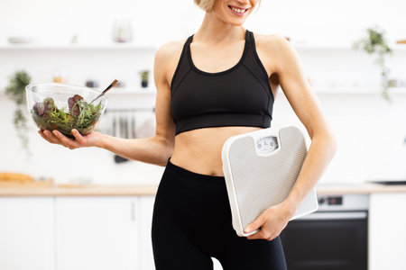 Fit woman holding salad bowl and bathroom scale in kitchenの写真素材