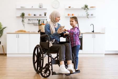 Muslim mother in wheelchair with daughter using mobile phone in kitchenの写真素材