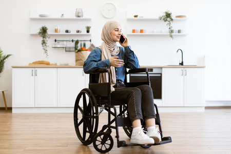 Muslim woman in wheelchair talking on phone at home kitchenの写真素材
