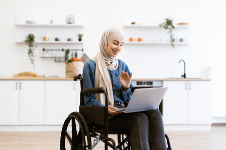 Muslim woman in wheelchair using laptop at home kitchenの写真素材