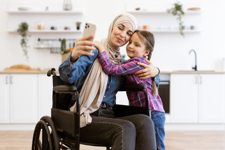Muslim mother in wheelchair taking selfie with daughter at homeの写真素材