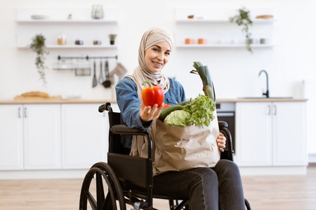 Muslim woman in wheelchair holding vegetables in modern kitchenの写真素材