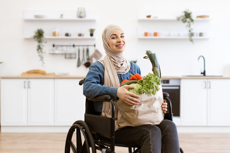 Muslim woman in wheelchair holding groceries in modern kitchenの写真素材
