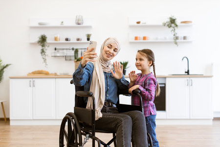 Muslim mother in wheelchair and daughter video calling together at homeの写真素材