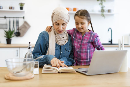 Muslim mother and daughter using laptop and recipe book in kitchenの写真素材