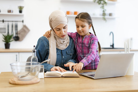 Muslim mother and daughter using laptop and recipe book in kitchenの写真素材