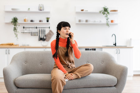 Young woman sitting on sofa talking on smartphone in cozy kitchenの写真素材
