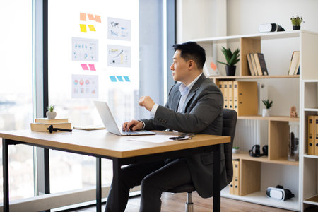 Asian businessman working on laptop in modern office settingの写真素材