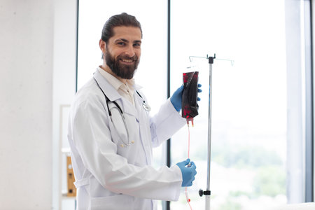 Male doctor with beard and long hair in white coat holds dropper with blood bag. He is preparing for transfusion in modern clinic. Doctor exudes confidence and professionalism.の写真素材