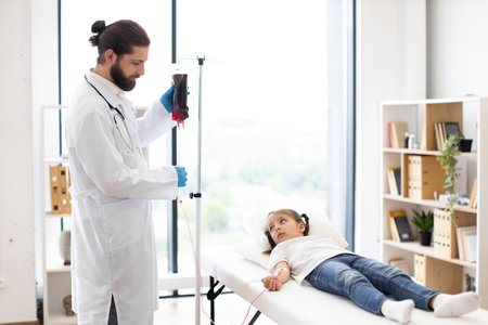 Male doctor providing blood transfusion to young girl lying on hospital bed. Medical professional wearing white coat, standing next to girl who appears calm.の写真素材