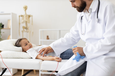 Male doctor gives blood transfusion to young Caucasian girl in hospital. Girl lies on bed with comfortable pillows, receiving medical attention. Doctor in lab coat and stethoscope ensures well-being.の写真素材