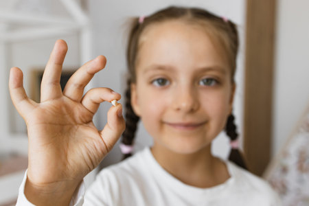 Little Caucasian girl smiling while holding recently lost baby tooth at home, symbolizing childhood...