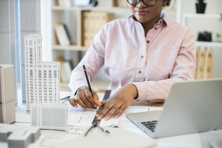 African woman architect focused on drawing city blueprint with scale models. Professional workspace includes laptop and drafting tools, highlighting creativity and precision in architectural design.の写真素材