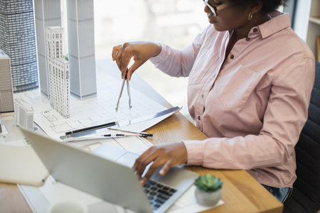 African woman architect using compass to measure dimensions on modern city project model. She is focused on design details at her workstation, surrounded by architecture tools and laptop.の写真素材