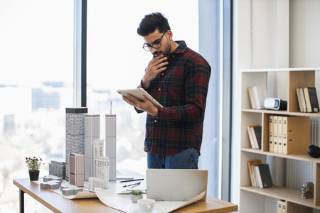 Indian male architect uses digital tablet to design modern city project. He stands in office near building models, displaying concentration and creativity.の写真素材