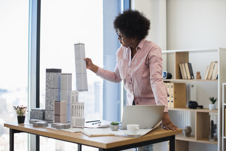 African American female architect in office setting holding building model. Young professional develops modern city project, focusing on urban planning and architectural design.の写真素材