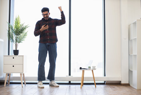Indian male in casual clothing celebrating success while looking at smartphone in bright modern room. Expresses joy and achievement.の写真素材