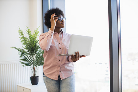 African American businesswoman standing near window holding laptop, wearing casual attire. Daylight and indoor plant create modern, productive atmosphere.の写真素材