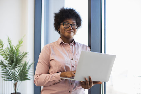 Young African American woman wearing glasses using laptop by window. She looks confident and...