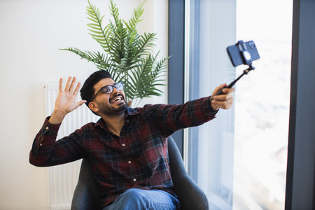Indian male in his 30s sitting and enjoying casual selfie moment with smartphone. Relaxed and...